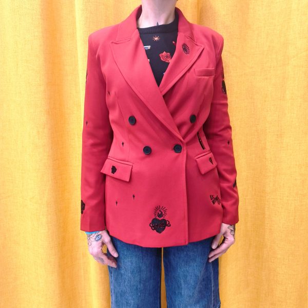 Blazer rouge épaules légèrement marquées. Motifs broderies noires (œil, cœur sacré, rouge à lèvres, diamant, revolver, rose, inscription " Ex-Wives Club). Tissu lisse et confortable, double rangée de boutons noires pour la fermeture, coupe ajustée.