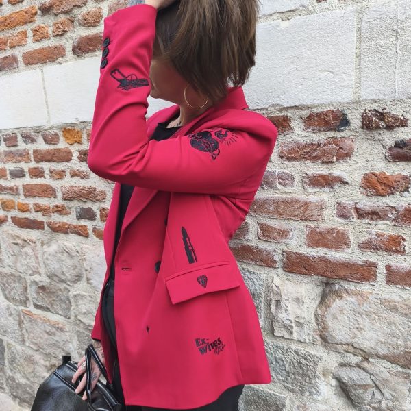 Blazer rouge épaules légèrement marquées. Motifs broderies noires (œil, cœur sacré, rouge à lèvres, diamant, revolver, rose, inscription " Ex-Wives Club). Tissu lisse et confortable, double rangée de boutons noires pour la fermeture, coupe ajustée.